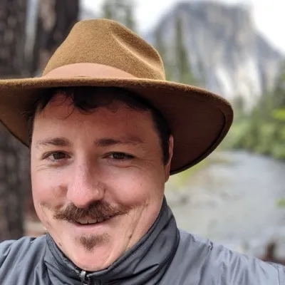 Kevin Kipp in Yosemite with a river with El Capitan in the background. He's smiling and wearing a tan hat.